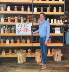 Photo of Richard Killian in a local shop. Richard Killian has managed complex budgeting and treasury operations, including annual budget preparation, cash-flow forecasting, bank reconciliation, public investments, and liquidity management. Photo has jars and canning including pickles in the background with Richard Killian standing in the front. A sign encourages people to shop local. 